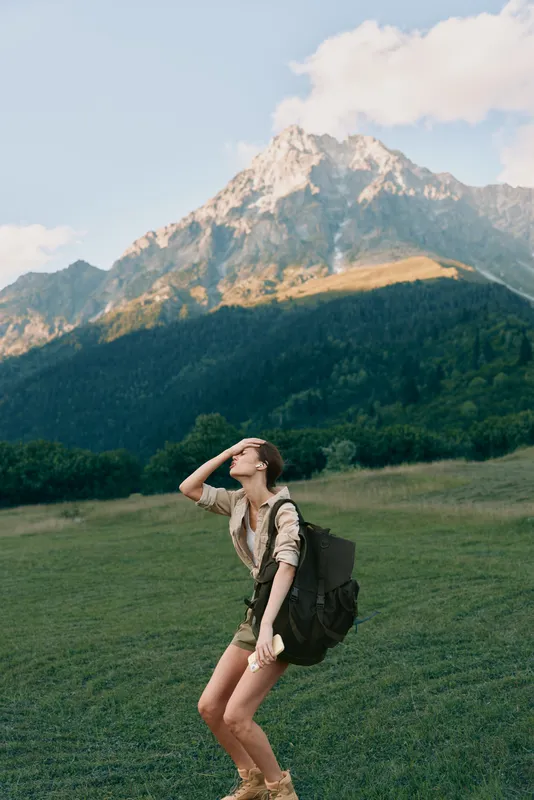 Cheerful woman outdoors, hiking in a sunlit meadow beneath mountains, with a backpack, relaxed posture, natural colors, fresh breeze, adventurous vibe with joyful energy