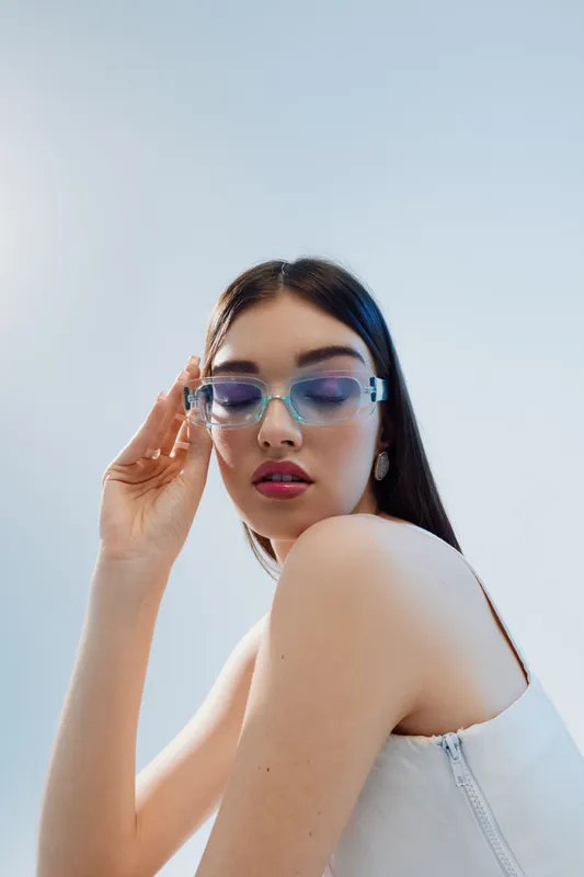 Young woman in white top wearing transparent sunglasses poses against a light background. Her makeup and hairstyle highlight beauty and fashion concepts.