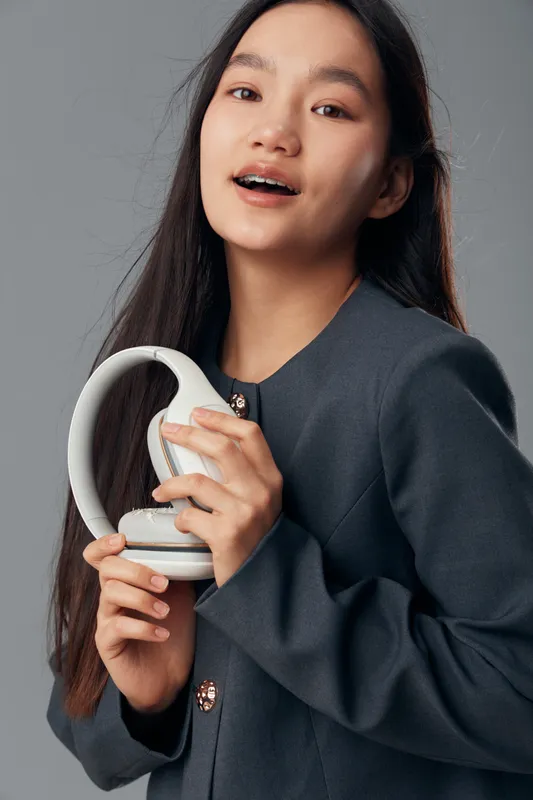 Young woman holding white headphones close to chest, smiling softly with long dark hair wearing dark blazer inside studio with neutral background and natural makeup.