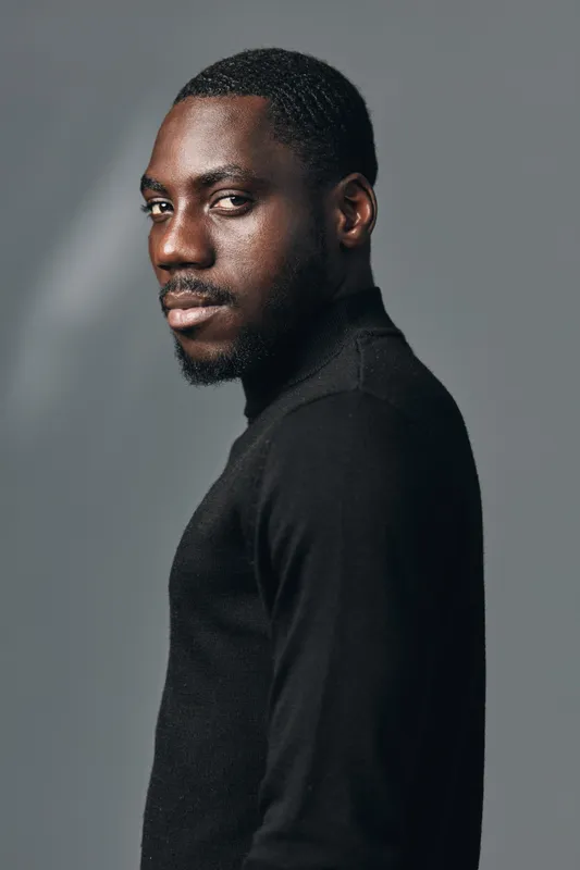 Portrait of African American man with dark skin wearing black turtleneck sweater in creative visual designer style. Studio shot with serious expression and side view on gray background.