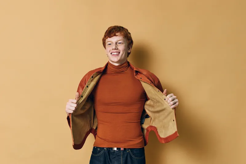 Young man with red hair smiling and wearing orange turtleneck and jacket standing against beige background. Casual style and happy expression with hands holding jacket open.