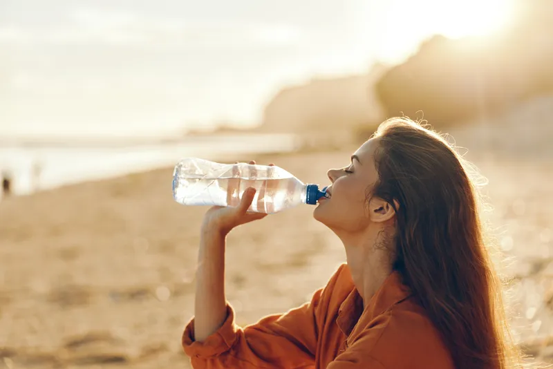 Woman drinking water outdoors on the beach during morning sunlight, hydration and wellness theme, she sips from a bottle, calm health focused self care, soft coastal breeze