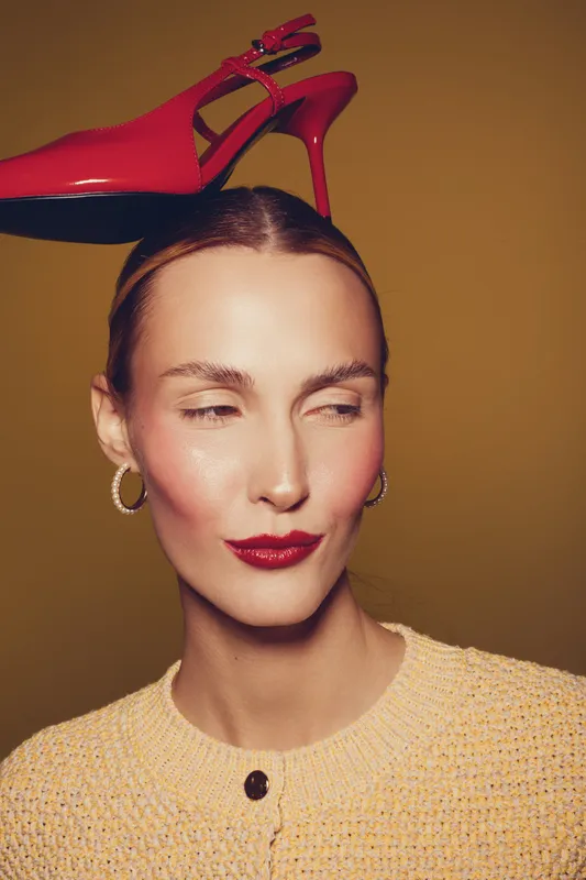 Portrait of a stylish woman with red lipstick and hoop earrings balancing a red high heel shoe on her head against a brown background. Concept of fashion, elegance, and creativity.