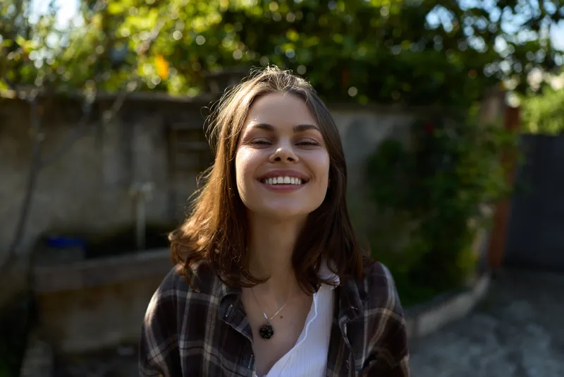 Bright smiling woman enjoys a sunny outdoor moment in a casual plaid shirt, light makeup, and simple jewelry, capturing a relaxed lifestyle portrait against a blurred garden backdrop