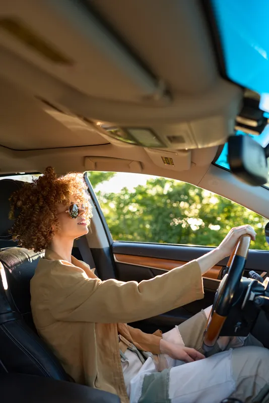 A woman with curly hair wearing retro sunglasses drives a car with warm filmic colors, portraying a relaxed lifestyle on the road and a casual daytime journey.