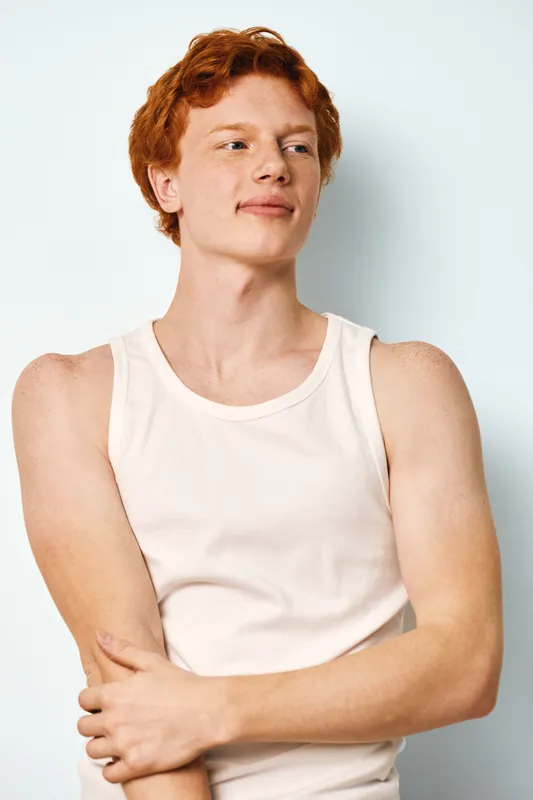 Young man with red hair and fair skin wearing white sleeveless shirt posing with crossed arms looking sideways against plain background in studio. Casual style and confident expression.