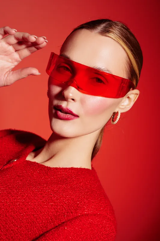 Stylish woman in red sweater and futuristic red sunglasses poses against red background. Concept of fashion, modern style, and bold color contrast in studio portrait photography.