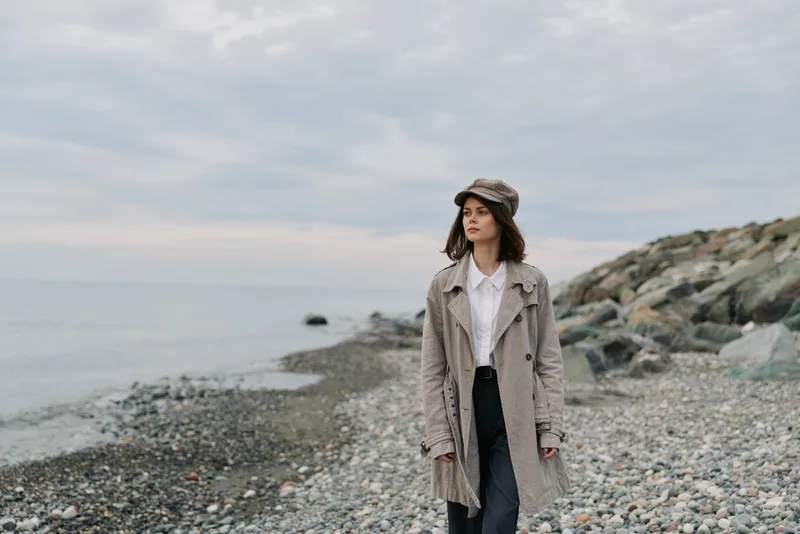 Young woman in warm coat and hat stands on rocky beach looking away. Overcast sky and calm sea in background create serene and contemplative mood outdoors.