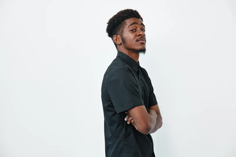 Young African American man with dark skin and short curly hair standing with arms crossed wearing black shirt on white background. Confident expression and casual style portrait.
