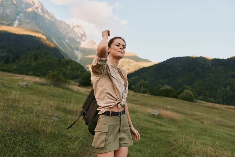 Smiling woman hiking outdoors with a backpack in a mountainous landscape, embracing nature and trail adventure on a sunny day, conveying exploration, wellness, and outdoor lifestyle.