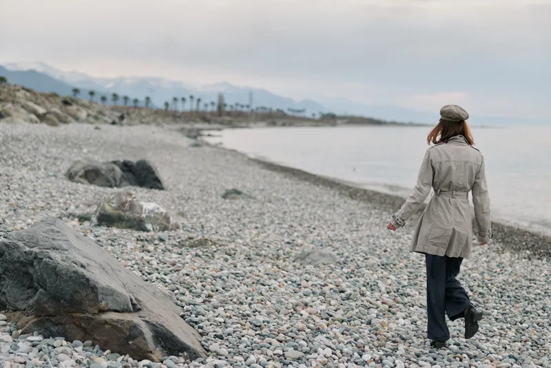Woman in beige coat and hat walks alone on a rocky beach by the sea under cloudy sky. Concept of solitude, nature, autumn, and peaceful outdoor lifestyle by calm water.