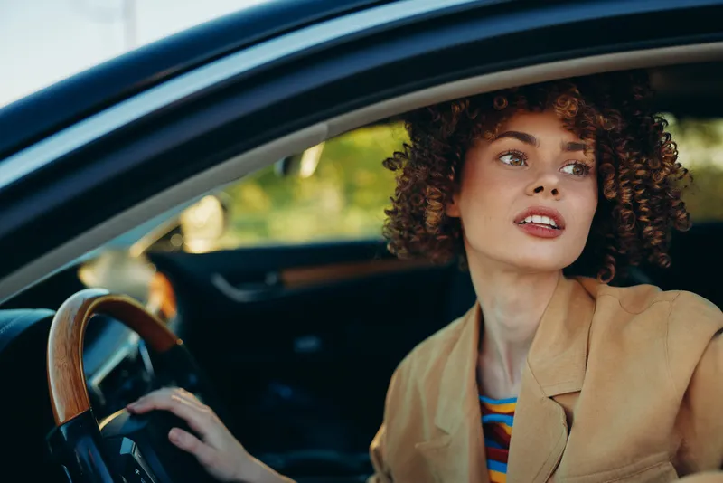 Bright woman in a car, smiling softly, retro vibe with warm sun, blazer and striped top, candid lifestyle moment, natural colors and lush light through the window