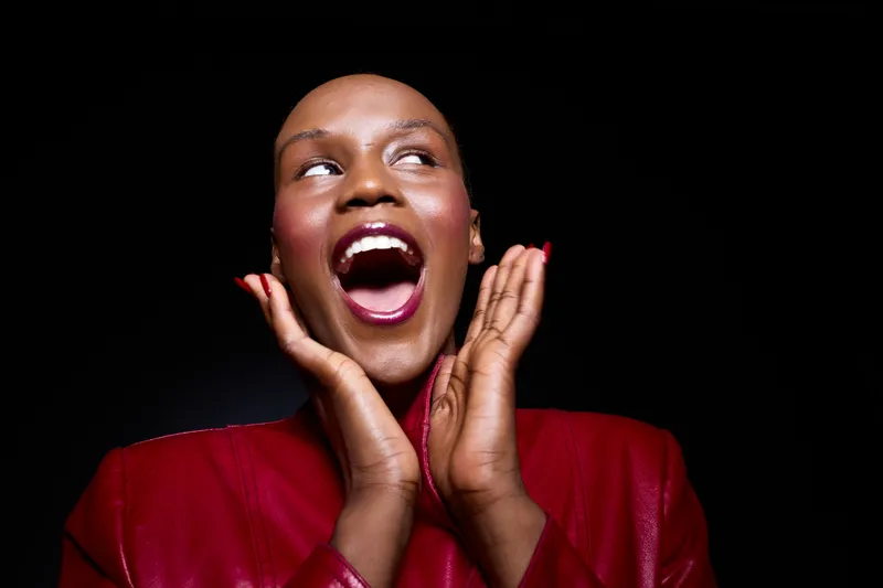 Excited happy black woman with dark skin wearing red jacket, smiling widely with open mouth and hands near face. Concept for joy, emotion, celebration, and positive expression on black background.