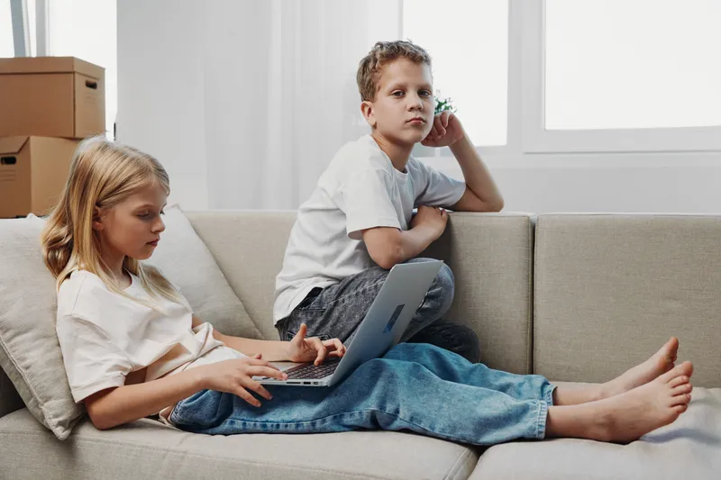 Two children spend time indoors on a sofa, one using a laptop while the other looks away, capturing a quiet domestic moment in a bright living room.