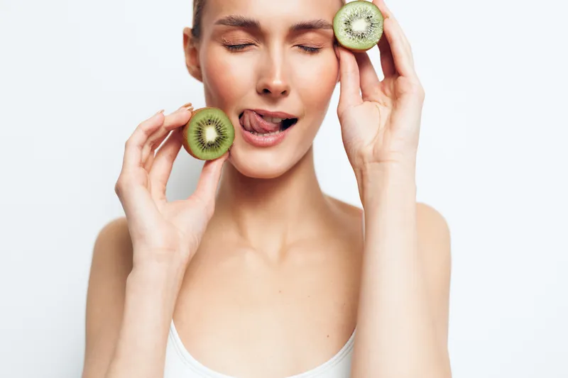 Young woman with a white tank top smiles softly while holding kiwi slices near her mouth against a clean white background, conveying a fresh skincare moment with playful charm.