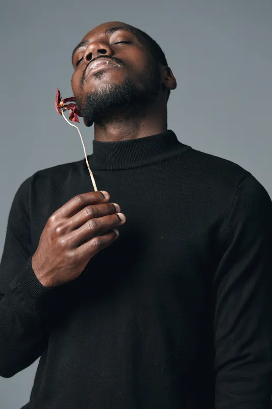 Black man with closed eyes holding dried flower near face in creative conceptual visual style. Portrait of African ethnicity person wearing black sweater on gray background.
