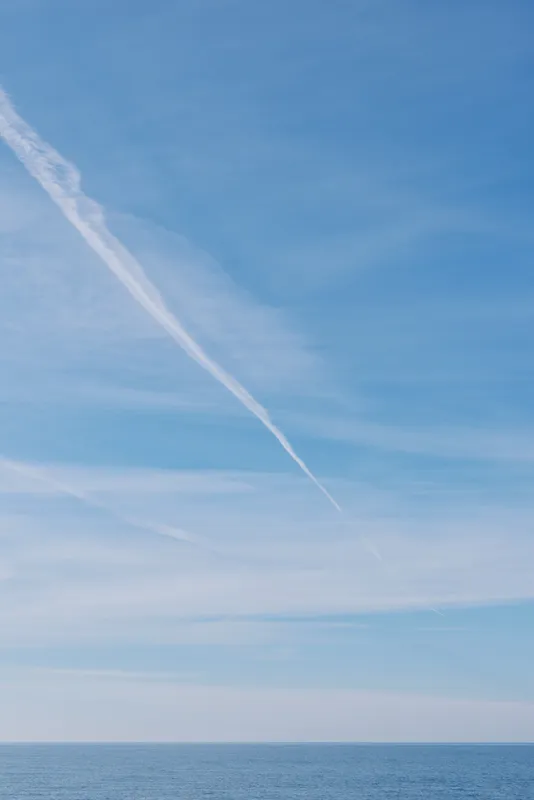 Blue sky above calm sea with distant horizon and a white contrail slicing through clouds, creating a tranquil landscape perfect for travel, nature, and serene inspiration.