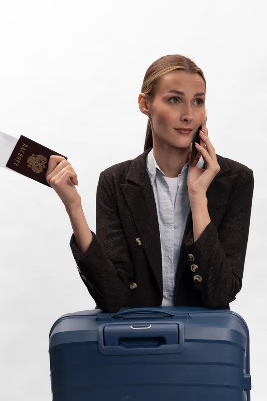 Young woman holding passport and talking on phone stands behind blue suitcase against white background. Concept of travel planning, trip preparation, and communication before journey.