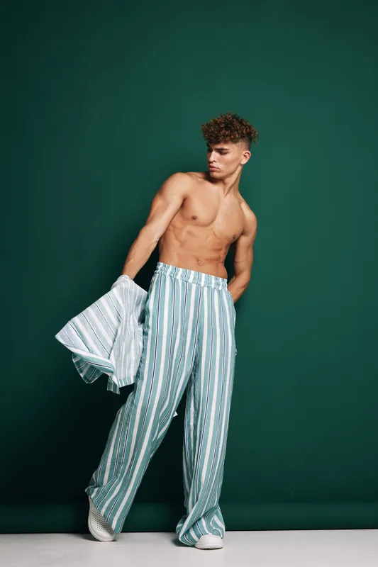 Young man with curly hair wearing striped wide pants and holding a matching shirt against green background. Fashion, casual style and confident pose in studio lighting.