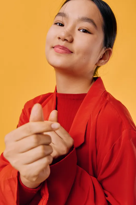 Portrait of confident woman in red clothes making finger heart gesture with smile on yellow background. Positive emotions, happiness, and cultural sign of love or appreciation closeup.