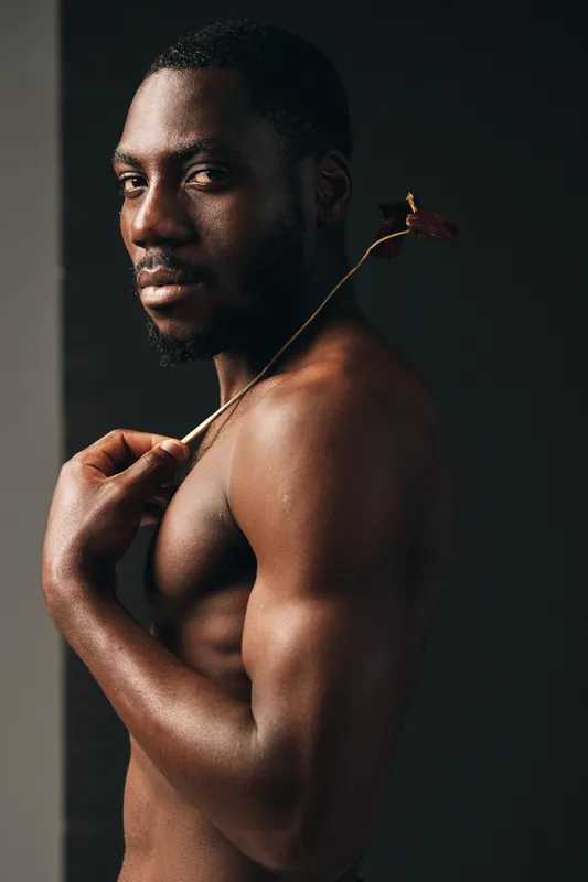 African man with muscular body and beard holding dried flower on shoulder in creative conceptual visual style with dark background and moody lighting for artistic portrait.