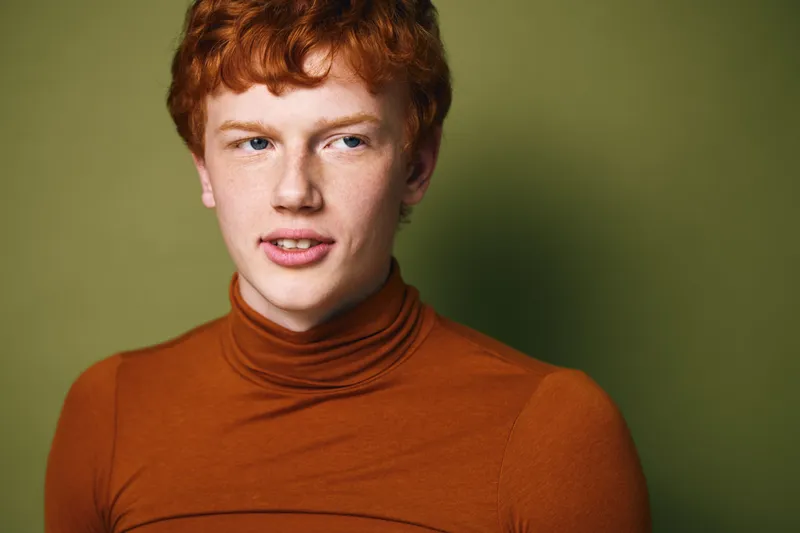 Young man with red hair and freckles wearing brown turtleneck sweater posing on green background with serious and thoughtful expression looking sideways away from camera.