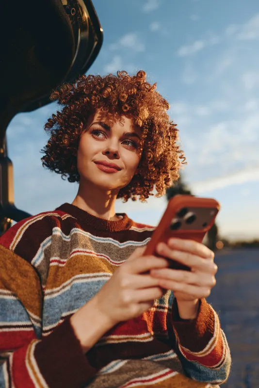 Woman with curly hair outdoors, holding a smartphone and showing a smile, Warm sunset light, colorful sweater, relaxed lifestyle mood, open sky and soft wind feel calm today