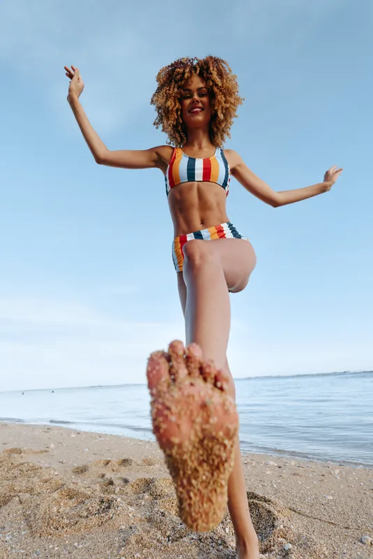 Young woman in colorful striped swimsuit standing on sandy beach, smiling joyfully, blue sky background, relaxed summer atmosphere, barefoot with sand on foot