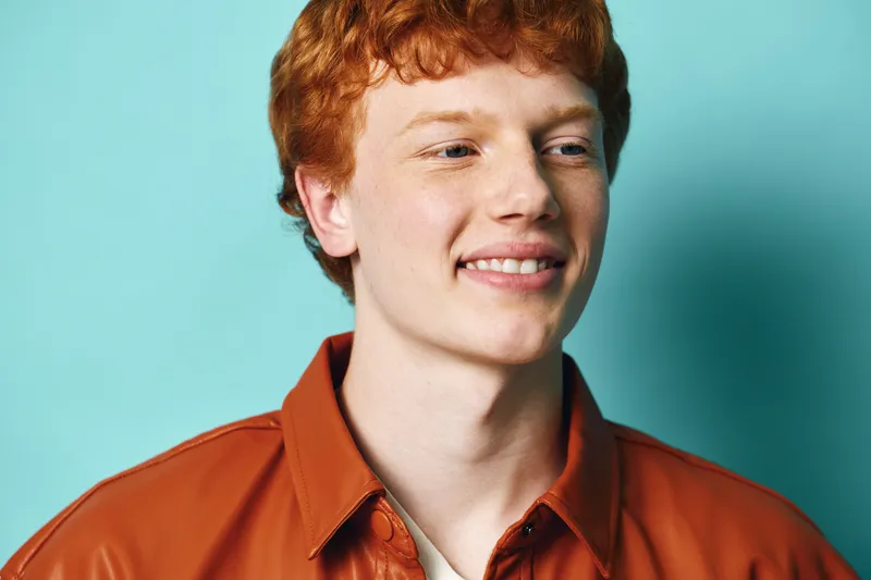 Young man with red hair smiling in casual orange shirt against blue background. Portrait of happy male teenager with freckles in studio lighting and relaxed expression.
