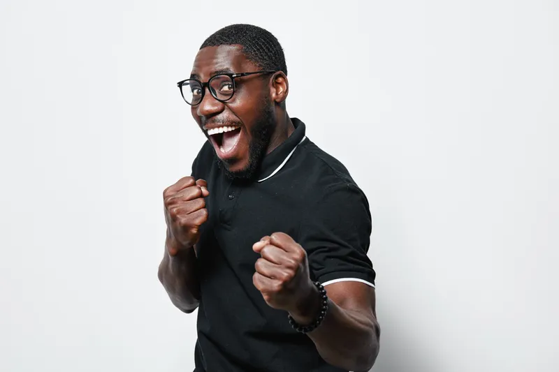 African American man with dark skin and glasses celebrating success with clenched fists and joyful expression wearing black polo shirt on white background in studio.