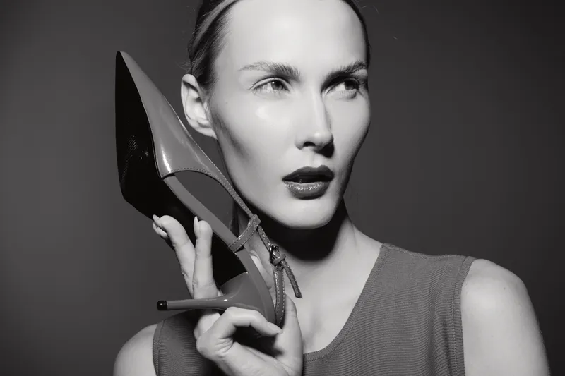 White woman with fair skin holds high heel shoe near face posing in studio. Concept of fashion, style, and elegance in monochrome.
