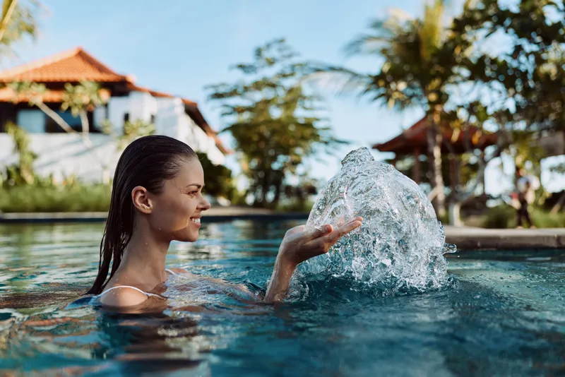 Cheerful woman in a sunny pool, laughing as she splashes water, relaxed pose, summer breeze, resort atmosphere, vibrant blue water, tropical backdrop, casual swimwear, carefree lifestyle moment