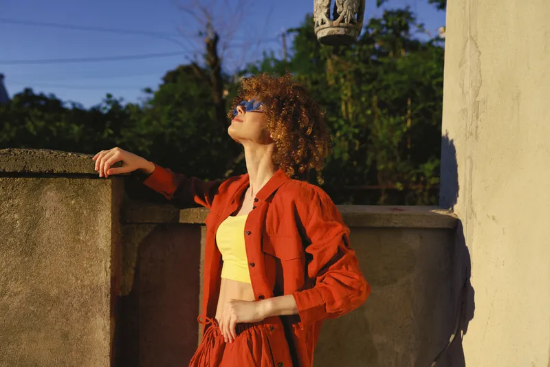 A young woman with curly hair leans against a sunlit stone railing, wearing a bright red outfit and oversized sunglasses, basking in warm afternoon light on an outdoor terrace