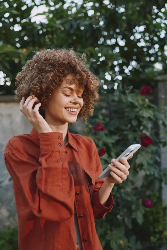 A smiling woman, lifestyle oriented, uses a phone in an outdoor garden with flowers and lush greenery, conveying casual, confident moments and everyday connectivity in a bright setting