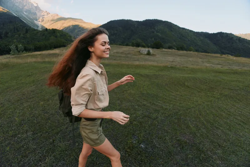 Smiling woman with backpack hiking through a grassy field under mountains. Outdoors lifestyle and nature scene highlight adventure, travel, and carefree mood.