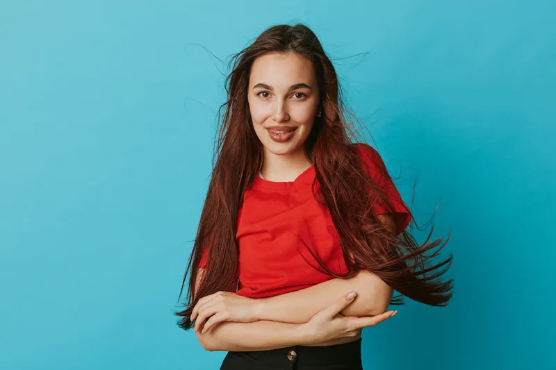 Young woman with long brown hair wearing red shirt stands with crossed arms smiling confidently on blue background. Casual style, portrait, positive expression and relaxed pose in studio.