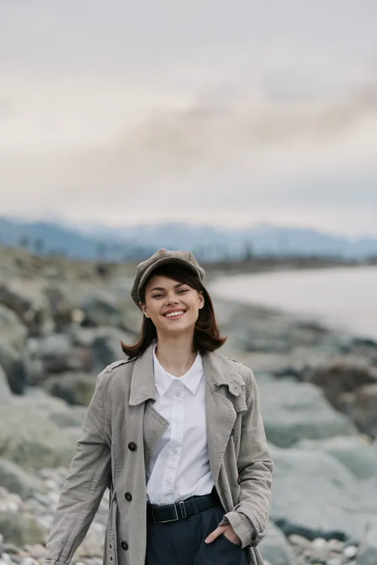 Smiling young woman in coat and cap stands on rocky beach with mountains and water in background. Overcast sky creates calm mood for outdoor lifestyle and nature concept.