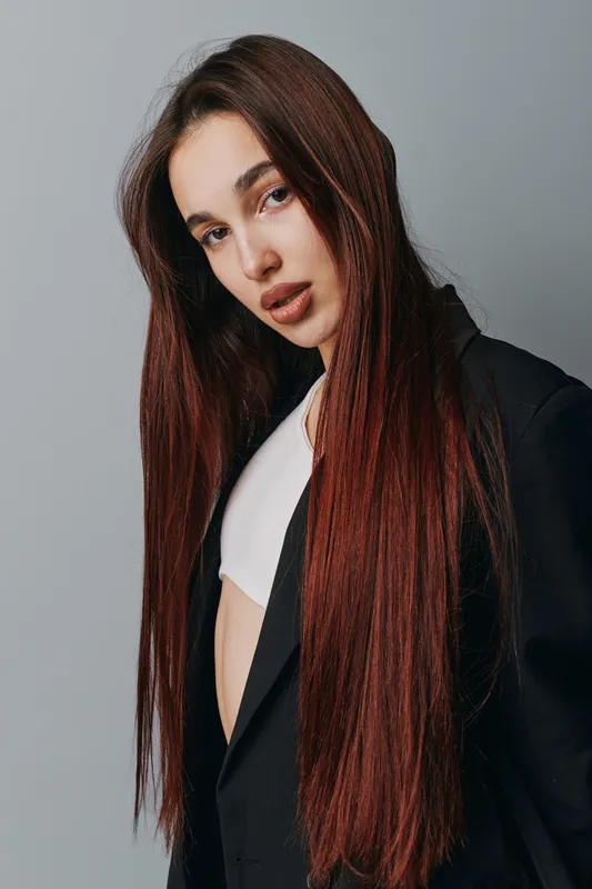 Young woman with long straight hair wearing black blazer and white top posing against gray background. Fashion portrait with natural makeup and confident expression for beauty and style.