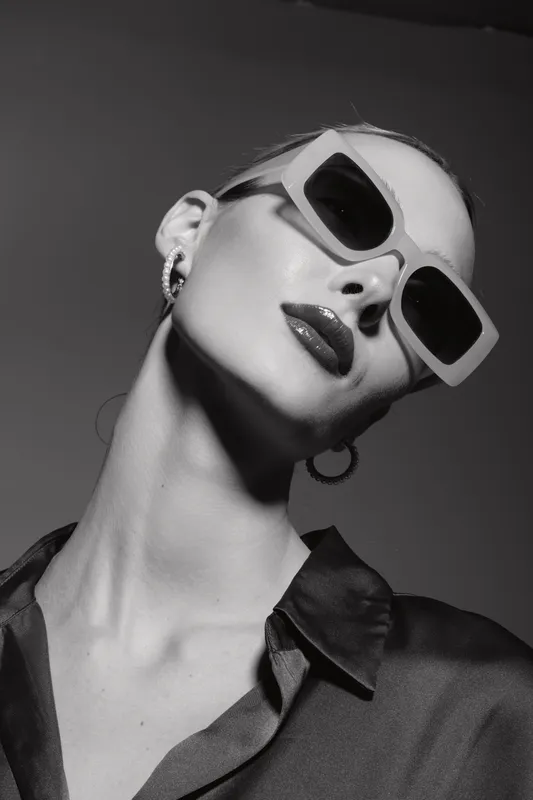 Stylish white woman with fair skin wearing oversized sunglasses and hoop earrings poses confidently against a dark background in a high fashion black and white portrait.