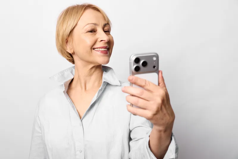 Woman selfie smartphone smiling portrait elderly blonde, senior person holds phone, cheerful expression, looking at camera, isolated on neutral background, home lifestyle