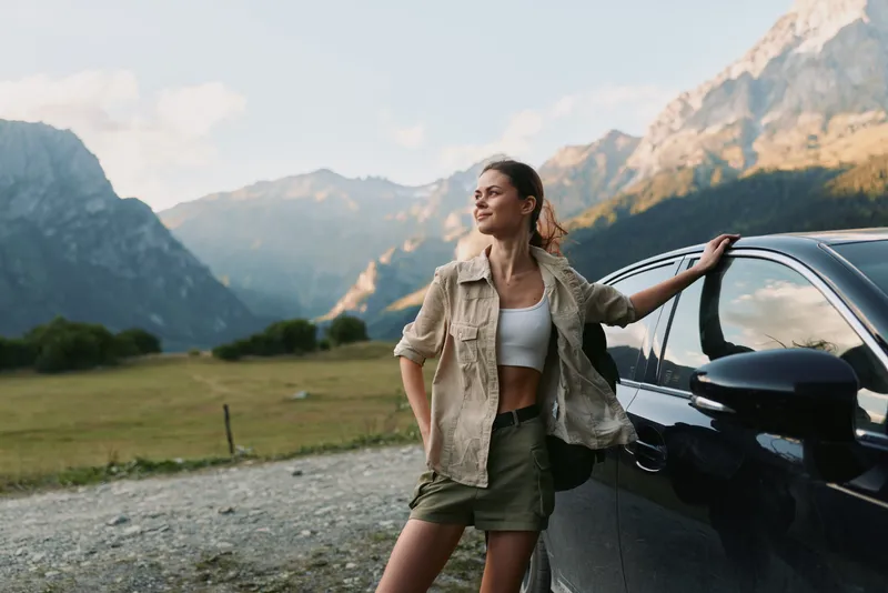 Woman posing beside a car in a mountain landscape, casual outfit, outdoorsy mood. Lifestyle scene highlighting freedom, travel, and nature exploration.