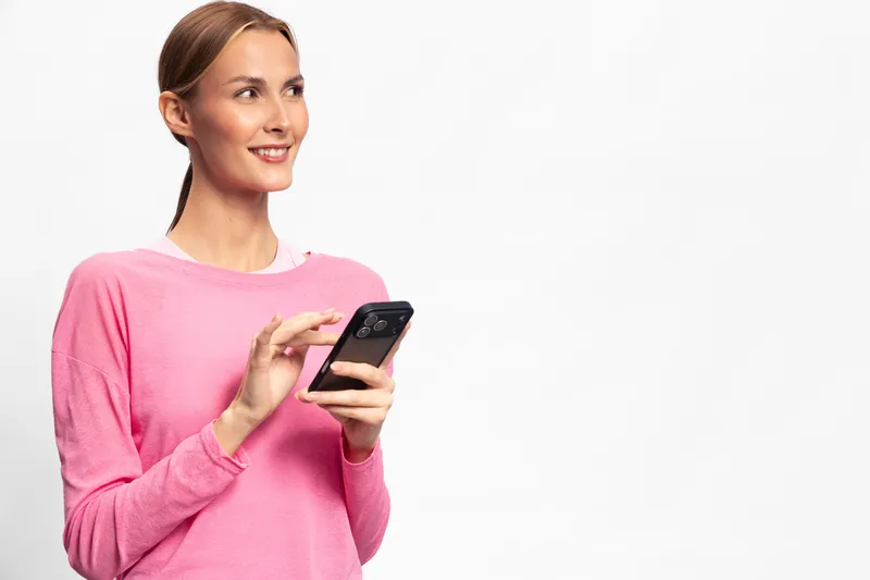 Young woman in pink sweater smiling while using smartphone on white background. Concept of communication, technology, lifestyle, and positive emotions in casual setting.