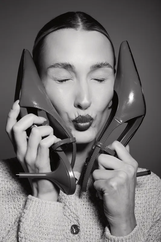 White woman with fair skin holding two high heel shoes near face, eyes closed, and puckering lips. Concept of fashion, style, and elegance in black and white.