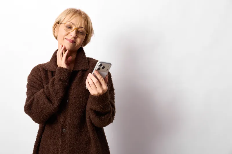 Elderly woman portrait indoors, holding smartphone near her face, brown coat, gentle smile, thoughtful gaze, white background, calm communication moment, soft studio light