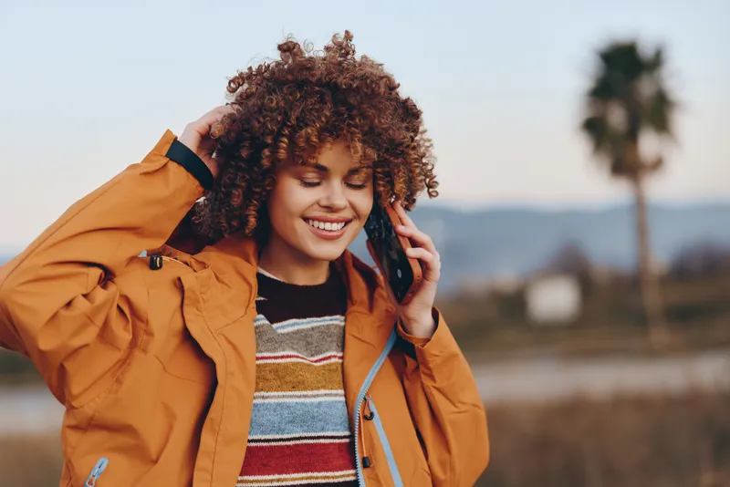 Woman lifestyle outdoors portrait, smiling curly hair woman in orange jacket adjusts hair, casual vacation mood, bright daylight, soft mountains background, blurred trees, open air