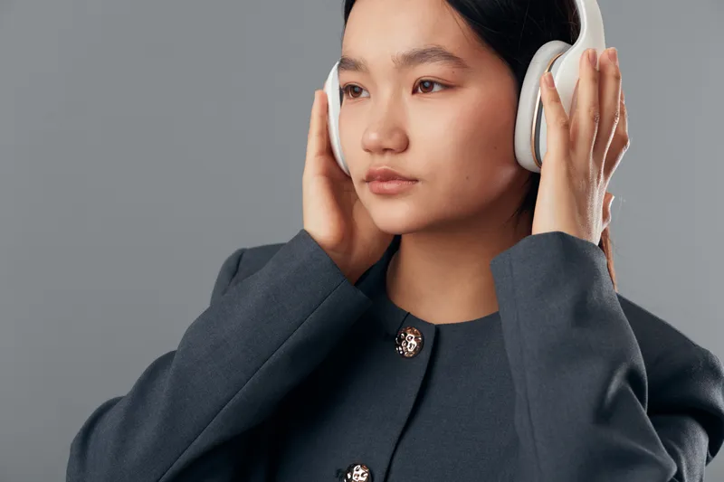 Woman wearing headphones and looking thoughtful with hands adjusting device over ears in studio environment. Female adult focusing on music or audio with gray background and casual clothing.