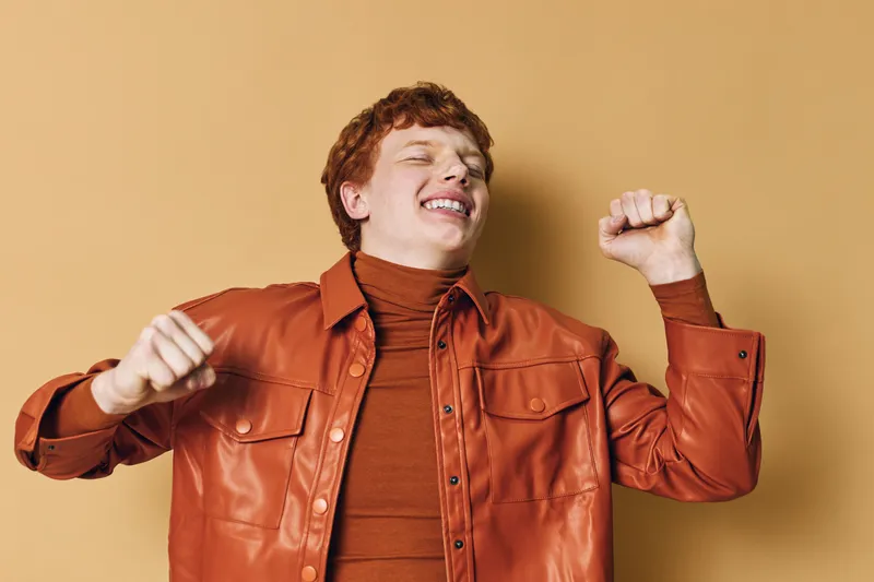Young man with red hair wearing brown leather jacket smiling with closed eyes and raised fists in celebration on beige background in studio.