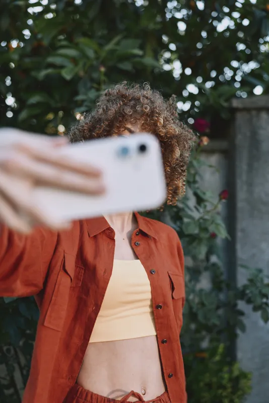 Woman taking a selfie outdoors, wearing a burnt orange jacket and crop top, cheerful expression, sunlit scene with lush greenery and relaxed lifestyle