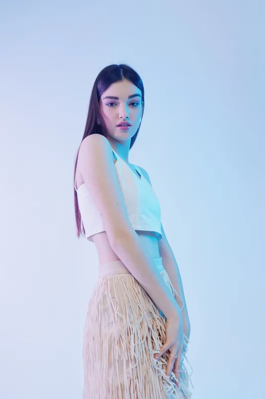 Young woman with long dark hair wearing a white crop top and fringed skirt poses against a light blue background. Concept for woman beauty fashion and modern style.