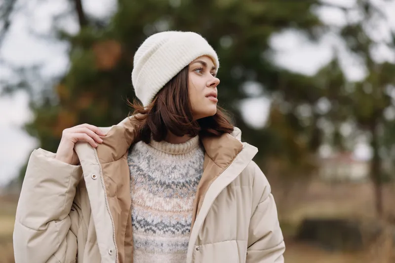 Woman in a beige puffer coat and white beanie stands outdoors, gazing to the side, showcasing winter fashion in a calm, natural portrait amid autumn scenery.